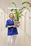 Helsearbeider i blå uniform står i sykehuskorridor og holder en stor grønn potteplante.